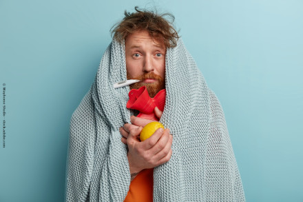 Ein Mann mit zerzaustem Haar und rotem Bart ist in eine graue Decke eingehüllt. Er hat ein Thermometer im Mund und hält ein rotes Stethoskop sowie eine Zitrone in seinen Händen. Der Hintergrund ist hellblau.