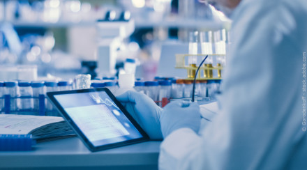 Ein Wissenschaftler in einer Laborumgebung untersucht Daten auf einem Tablet. Im Hintergrund sind mehrere Reagenzgläser, Laborgeräte und Probenbehälter zu sehen. Der Wissenschaftler trägt einen weißen Laborkittel und blaue Handschuhe.