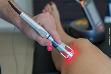Nahaufnahme eines Physiotherapeuten, der eine Lasertherapie-Einheit auf ein Knie anwendet. Der Laser leuchtet rot und wird auf die Haut gerichtet.