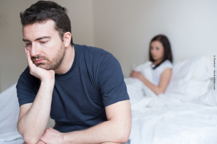 Ein Mann mit dunklem Haar und Bart sitzt nachdenklich auf einem Bett, stützt seinen Kopf auf seine Hand und schaut nach unten. Im Hintergrund sitzt eine Frau mit dunklem Haar in einem weißen Oberteil auf dem Bett, sie wirkt ebenfalls nachdenklich.