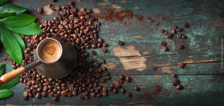 Ein traditionelles Kaffeekännchen, gefüllt mit heißem Kaffee, steht auf einem Tisch, der mit verstreuten Kaffeebohnen und gemahlenem Kaffee bedeckt ist. Daneben sind grüne Blätter zu sehen.
