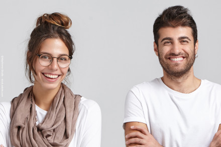 Ein Mann und eine Frau stehen vor einem grauen Hintergrund und lächeln in die Kamera. Die Frau trägt eine Brille und einen beigen Schal. Der Mann hat dunkles Haar und trägt ein weißes T-Shirt.