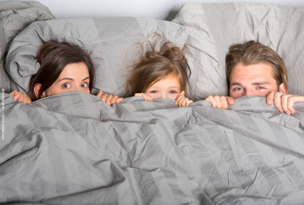 Eine Frau, ein Kind und ein Mann liegen im Bett und schauen überrascht über die Bettdecke, die sie bis zur Nase hochgezogen haben.