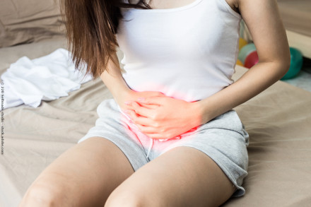 Eine Frau sitzt auf einem Bett und hält ihren schmerzenden Unterbauch, der rot hervorgehoben ist. Sie trägt ein weißes Tanktop und graue Shorts. Im Hintergrund sind bunte Objekte und ein weißes Kleidungsstück auf dem Bett zu sehen.