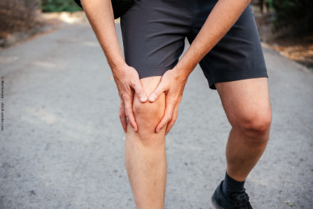 Eine Person in kurzer Hose legt ihre Hände um ihr schmerzendes Knie während eines Spaziergangs auf einem Weg.