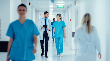 Mehrere Ärzte und Pflegekräfte gehen einen hell erleuchteten Krankenhausflur entlang. Ein Arzt in weißem Kittel und eine Krankenschwester in blauer Uniform führen ein Gespräch, während sie gehen. Es sind weitere medizinische Fachkräfte zu sehen.
