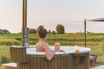 Eine Frau sitzt in einem Whirlpool im Freien mit Blick auf eine grüne Landschaft. Sie trägt ein türkisfarbenes Bikinioberteil und hält ein Glas Orangensaft in der rechten Hand. Ein weiteres Glas steht am Rand des Whirlpools.