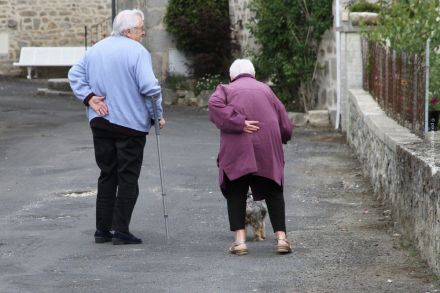 Ein älteres Paar geht langsamen Schrittes auf einer schmalen Straße. Die Frau trägt eine lila Jacke und der Mann eine hellblaue Jacke und führt einen Gehstock. Beide haben eine Hand auf ihren unteren Rücken gelegt, als hätten sie Rückenschmerzen.