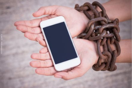 Nahaufnahme von Händen, die ein Smartphone halten, während sie von dicken, rostigen Ketten gefesselt sind.