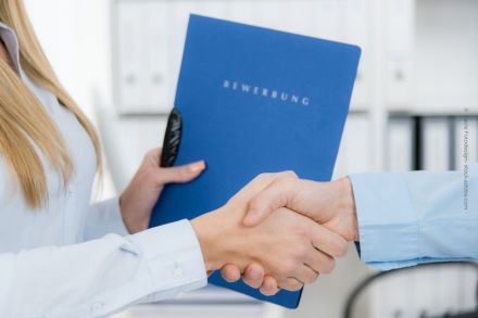 Person in weißem Hemd, die einer anderen Person in blauem Hemd die Hand schüttelt. Die Person im weißen Hemd hält eine blaue Bewerbungsmappe mit dem Wort "BEWERBUNG" darauf. Im Hintergrund sind unscharfe, weiße Büromöbel zu sehen.