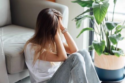 Eine Frau sitzt auf dem Boden, lehnt sich gegen ein Sofa und hält ihren Kopf in ihren Händen. Neben ihr steht eine grüne Zimmerpflanze. Die Szene wirkt nachdenklich und melancholisch.