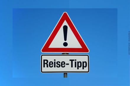 Ein dreieckiges Verkehrsschild mit rotem Rand und schwarzem Ausrufezeichen auf weißem Hintergrund. Darunter ein rechteckiges Schild mit der Aufschrift "Reise-Tipp" in schwarzer Schrift. Der Hintergrund ist ein klarer blauer Himmel.