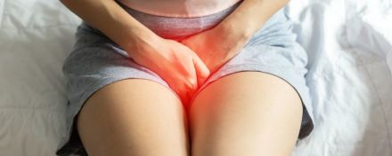 Eine Person sitzt auf einem Bett und hält sich den schmerzenden Unterbauch, der rot hervorgehoben ist. 