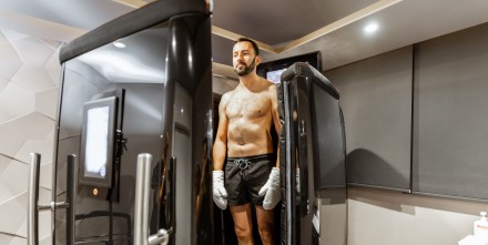 Ein Mann in Shorts und Handschuhen steht in einer Kryokammer in einem modernen Wellness-Zentrum. Er bereitet sich auf eine Kältetherapie vor, umgeben von futuristischer Ausrüstung und gedämpfter Beleuchtung.