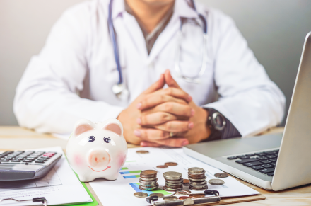Arzt sitzt mit gefalteten Händen am Schreibtisch. Vor ihm ein Taschenrechner, Sparschwein, Münzen und ein Laptop.