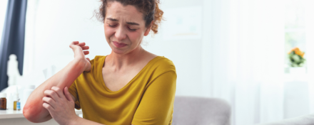 Frau in gelbem Oberteil kratzt sich am geröteten Unterarm, sichtbar von Hautreizungen betroffen. Das Bild zeigt typische Symptome von Neurodermitis, wie Rötungen und Juckreiz. Im Hintergrund ein heller Wohnraum mit Pflanzen und Pflegeprodukten.