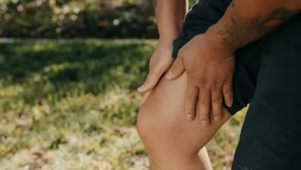 Eine Person umfasst ihr schmerzendes Knie mit beiden Händen. Es wird der Eindruck von Gelenk- und Muskelschmerzen, nach einer körperlichen Aktivität vermittelt. Der Hintergrund ist unscharf und zeigt eine Wiese im Sonnenlicht.