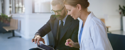Ärztin im weißen Kittel bespricht mit einem Anzugträger am Tablet digitale Marketingstrategien zur Patientenakquise in einer modernen Praxisumgebung.