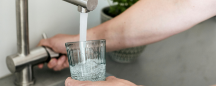 Eine Person füllt ein Glas mit Leitungswasser an einem Wasserhahn in der Küche – Symbolbild für Trinkwasserhygiene und Schutz vor Legionellen im Haushalt.