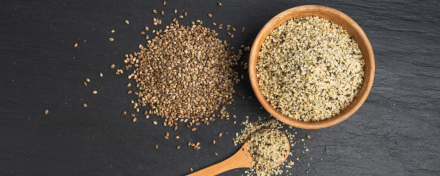 Ungeschälte Hanfsamen und geschälte Hanfkerne in einer Holzschale sowie auf einem Löffel, arrangiert auf dunkler Steinoberfläche – Darstellung verschiedener Formen von Hanfsamen für Ernährung und Lebensmittelverarbeitung.