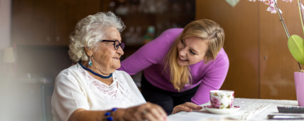 Ältere Frau und jüngere Betreuungsperson sitzen gemeinsam am Tisch, lächeln sich an und führen ein Gespräch in einer wohnlichen Umgebung; eine Tasse Tee und eine Orchidee stehen daneben.