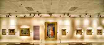 An art gallery with paintings on a cream-colored wall, dimly lit by overhead spotlights. Two wooden benches are placed in the foreground on a polished wooden floor. A large framed painting is centrally displayed.