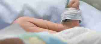 Close-up of a baby's foot with a medical sensor attached to it, lying on a white sheet in a hospital. The tiny foot has a bandage securing the sensor, indicating the baby is under medical care. Soft lighting gives a calm atmosphere to the scene.