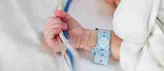 A close-up of a newborn baby lying on a white blanket. The baby is wearing a white outfit and a blue hospital identification bracelet on its wrist. The baby is holding a clear tube, and the image emphasizes the infant's small hand and arm.