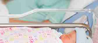 A newborn baby wearing a blue hat sleeps in a hospital bassinet, wrapped in a colorful blanket. In the blurred background, an adult lies in a hospital bed.
