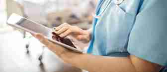 A healthcare professional in blue scrubs uses a tablet, with a stethoscope around their neck. A patient lies in a hospital bed in the blurred background, indicating a medical setting.