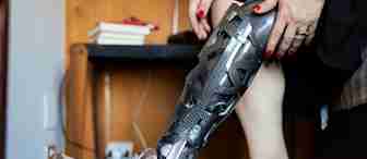 A person wearing sneakers is putting on a patterned, prosthetic leg. The person’s hands are adjusting the prosthesis near the knee. A wooden table with books and an electrical outlet are visible in the background.