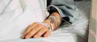 A person lying in a hospital bed with an intravenous (IV) line inserted in their hand, covered by a light blanket, suggesting a medical treatment or hospitalization.