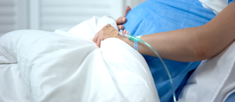 A pregnant woman wearing a blue hospital gown lies in a hospital bed with an IV drip attached to her hand. Her hands rest on her large belly, indicating she is in the later stages of pregnancy. The background shows white shutters.