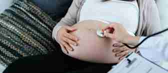 A pregnant woman sits on a sofa, holding her belly. A healthcare professional, whose face is not visible, uses a stethoscope to check the woman’s abdomen. A clipboard with medical documents is in the healthcare professional's hand. Pillows are in the background.