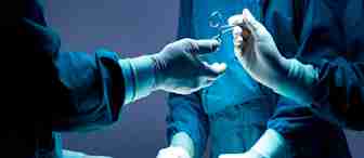 Surgeons in blue scrubs and gloves performing surgery under bright lights. One surgeon holds a pair of scissors while another assists with tools. The patient is draped with a blue surgical cloth.