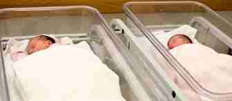 Two newborn babies are sleeping in separate hospital bassinets covered with white blankets. The bassinets are placed side by side in a calm, well-lit room. The babies are swaddled and appear peaceful.
