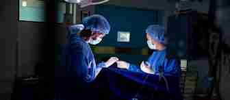 Two surgeons in blue scrubs and surgical masks work under bright lights in an operating room. One is performing a procedure while the other assists. Medical equipment is visible around them, and the background is dimly lit but identifiable as a medical facility.