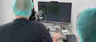 Two individuals in a laboratory setting are focused on a computer screen displaying a microscopic image. They are wearing surgical caps and gowns, indicating a sterile environment. The one on the left is manipulating a device, likely part of the microscope setup.