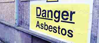 A large yellow sign on a boarded-up wall reads "Danger Asbestos" in bold black letters, indicating the presence of hazardous material. The wall and sign show some wear, suggesting an older, possibly abandoned building. Dry leaves are scattered on the ground nearby.