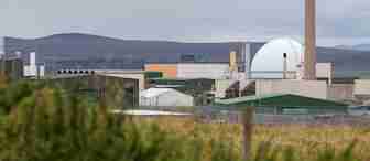 Industrial facility with a large dome-shaped structure and various buildings under a cloudy sky. In the foreground, there are grassy fields and fencing. Hills are visible in the background.
