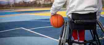 A person in a wheelchair holding a basketball sits on an outdoor court, ready to play. The court is painted blue and yellow, with clear sky and buildings in the background.