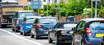 Several cars are lined up in a traffic jam on a city street during the daytime; the vehicles are stopped close together, with houses and greenery visible in the background.
