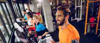 People exercise on treadmills in a well-lit gym. In the foreground, a man with a beard, wearing a bright orange shirt and earphones, runs on a treadmill with a phone strapped to his arm. Two people are visible in the background, also using treadmills.