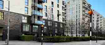 A city street with a zebra crossing leading to a modern apartment building. The buildings are surrounded by greenery and bare trees. The sky is clear and blue, and the streetlights are painted with black and white stripes and orange globes.