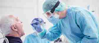 A healthcare worker in full protective gear, including a face shield, mask, and gown, swabs the mouth of an older patient seated in a clinical setting. Another healthcare worker in similar protective gear is seen in the background.