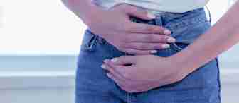 A person in a white shirt and blue jeans is shown from the waist down, holding their lower abdomen with both hands. The person's fingernails are manicured with light-colored nail polish. The background appears to be a bright indoor setting.