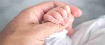 Close-up image of an adult hand gently holding a newborn baby's hand. Both hands are positioned against a soft, out-of-focus background, creating a tender and intimate moment of connection and care.