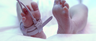 A close-up image of a newborn baby's feet. One foot is fitted with a medical pulse oximeter sensor, which has wires attached to it. The baby is lying down on a white blanket, with the image conveying a soft, tender moment.