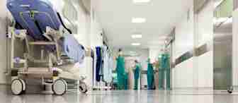 A hospital hallway with a patient bed on the left and medical staff in scrubs standing and talking in the background. The corridor is brightly lit with modern medical equipment and doors along the walls. The scene appears busy but organized.