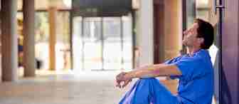 A person in blue scrubs and brown shoes is sitting on the floor, resting their back against a wall with eyes closed, appearing tired or contemplative. The background is out of focus, showing a well-lit hallway with large windows.
